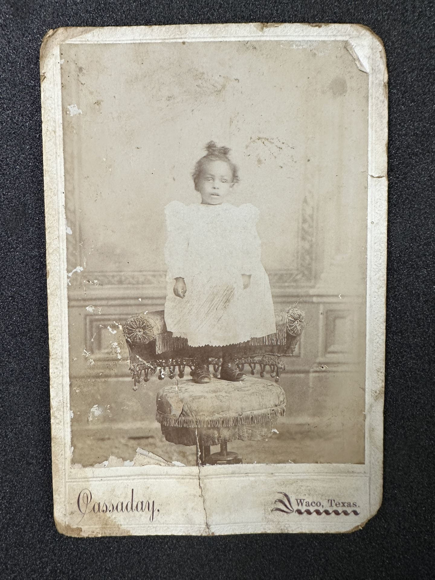 Black-and-white photograph of a young child in a white dress standing on a posing chair.