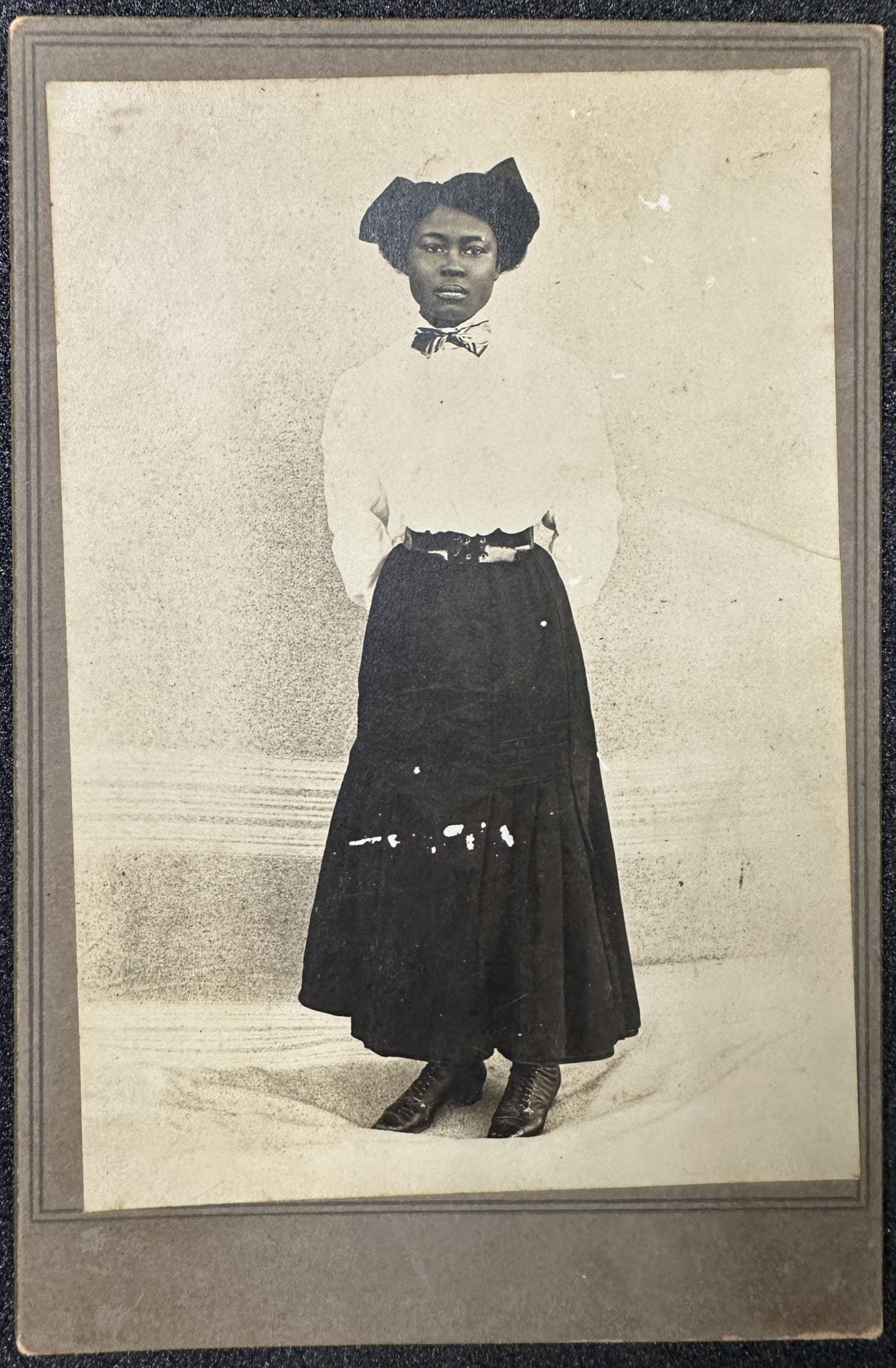Portrait of a standing Black woman wearing a white shirt and long dark trouser skirt.