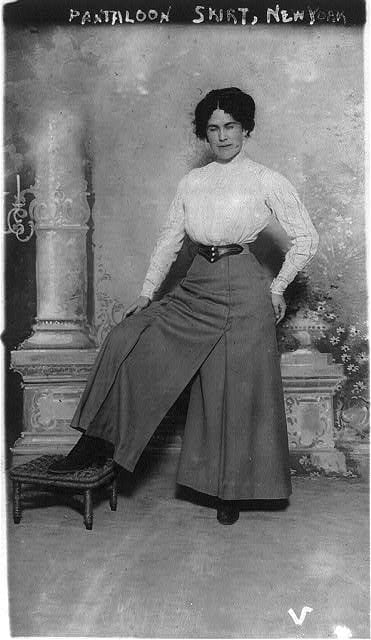Portrait of a white woman wearing a white shirt and dark trouser skirt standing with her right foot on a stool.
