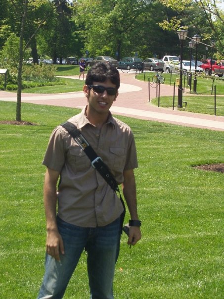 Person with dark hair and sunglasses standing in front of bright green grass and brick walkway
