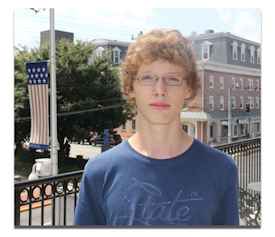 Person with curly red hair and glasses standing in front of a brick building.