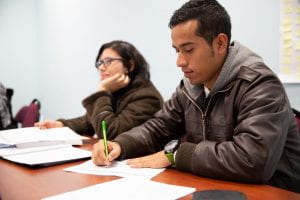 An ELI student in a leather jacket takes notes while another student looks up as if listening to the instructor.