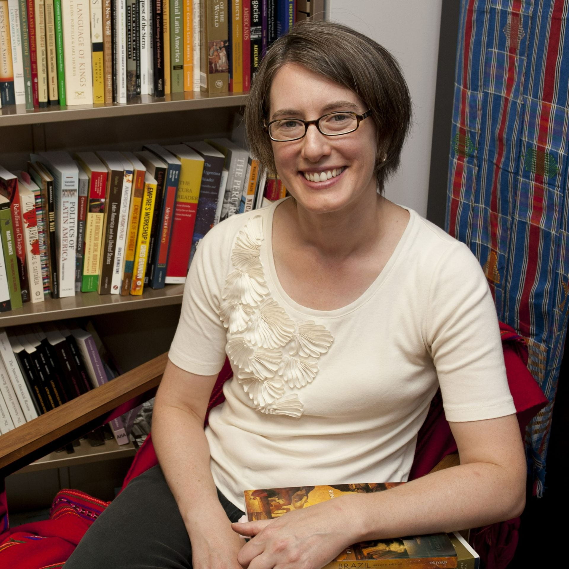 Prof. Buckley, a white, middle-aged woman with brown hair and glasses, wearing a short-sleeved white shirt, sits in a chair holding several books, with a full bookshelf and a colorful textile in the background.