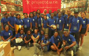 UD Fellows stand and sit for a group photo in front of a Project C.U.R.E. sign. 