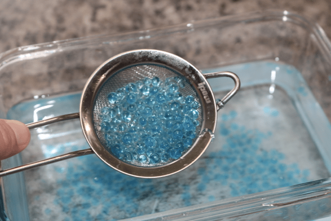 A photograph of a hand holding a wire mesh strainer filled with blue boba balls. The boba balls formed in the cloudy white solution of calcium chloride after sodium alginate solution was dropped into it. There are a few blue boba balls still left behind in the calcium chloride solution that were not scooped up by the strainer.