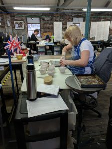 A woman sits at her workbench as she rubs the paper transfer print onto a teacup. She is holding the teacup in her left hand and a bristled brush in her right. Several teacups sit on top of her workbench.