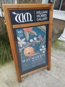 A sandwich board museum sign outside the William Morris Gallery in London, England with exhibition information including dates of the show, 7 October 2017-28 January, 2018, and an image of a May Morris embroidery with oranges and white blossoms on branches with multi-colored leaves on a teal green background.