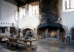 A large fireplace dominates the room, with blackness from fires reaching the ceiling. The blackness stands out against the white walls of the rest of the room. In the foreground sits a long table with cauldrons, bowls, and baskets of fresh vegetables.
