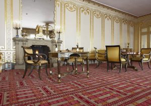 View of the corner of a large room with high ceilings and white walls painted with gilded decorative accents. Empire-style tables and chairs upholstered in yellow fabric stand atop a pink, gray and red carpet with a geometric pattern.