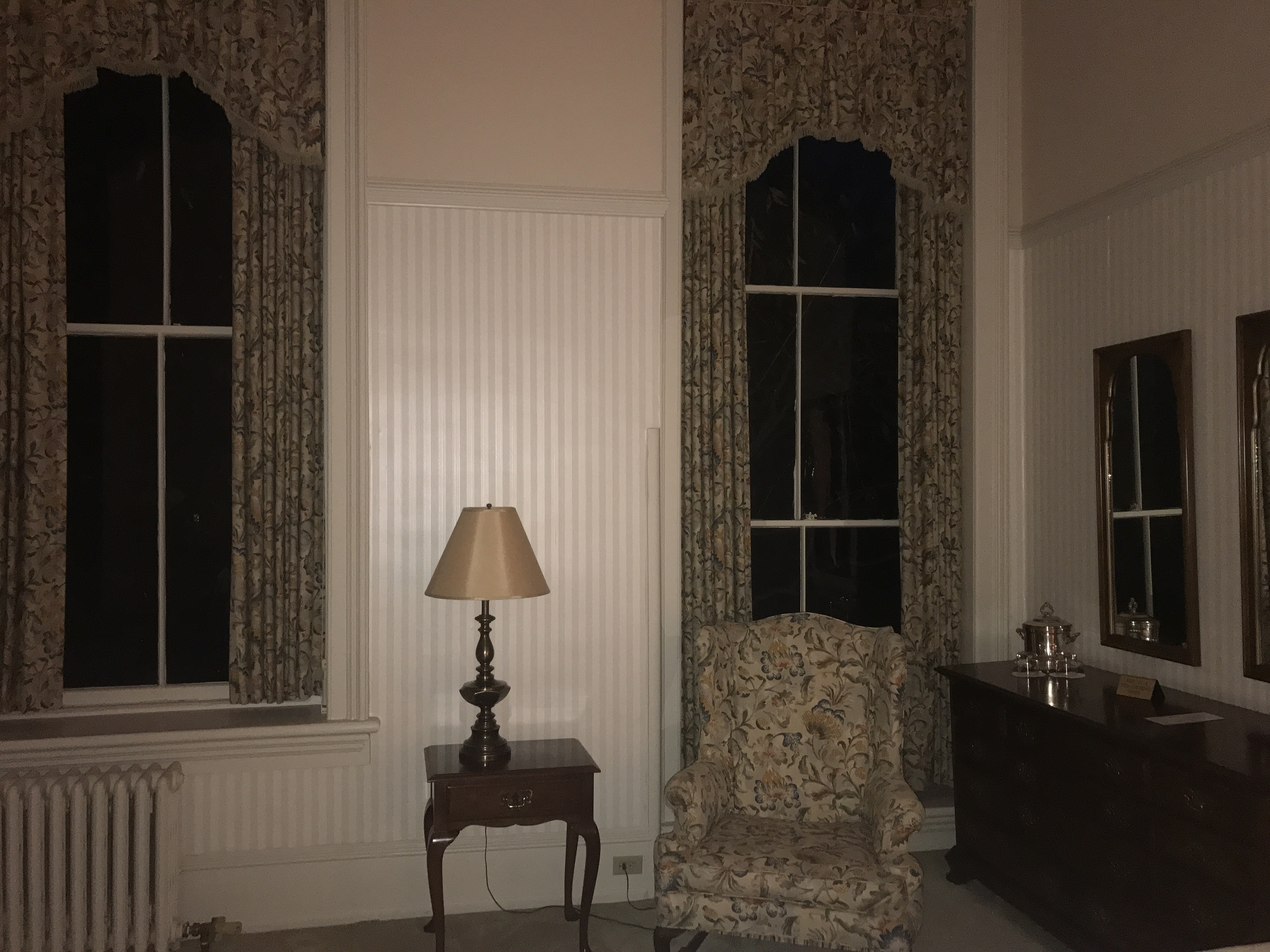 Oneida Room – A bedroom available for overnight stay in the Mansion House features very high ceilings, floral curtains, striped wallpaper, and an easy chair.