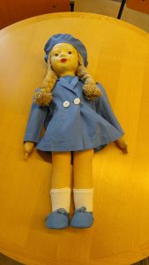 A doll with pale face and blonde braids is laid on a tabletop wearing a light blue coat with two white buttons, matching blue cap and shoes, and white calf-high socks.