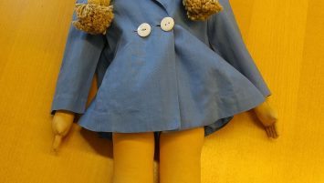 A doll with pale face and blonde braids is laid on a tabletop wearing a light blue coat with two white buttons, matching blue cap and shoes, and white calf-high socks.