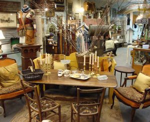 Interior of the antique store. Rectangular table in the center with two upholstered chairs on each side, two wooden chairs in the front. Many candlesticks and small cups sit atop table.