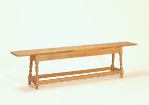 2.Wooden form on white background. The form is comprised of four turned legs with connecting rails. The seat rests atop the legs and has a significant overhang.
