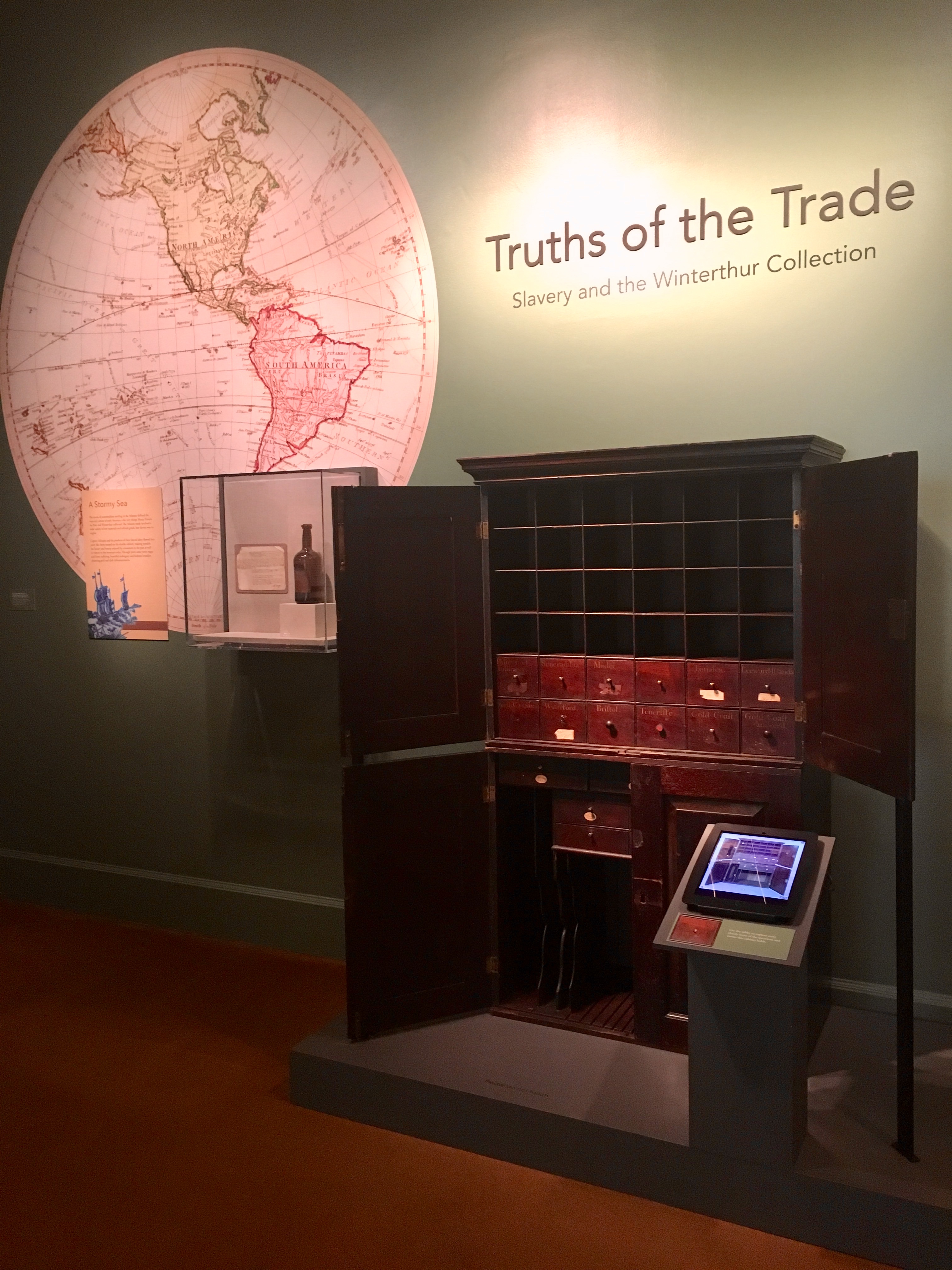 A view inside the Truths of the Trade exhibit featuring the mahogany double cabinet at center along with the the ipad touch screen interactive in the front.
