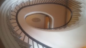 Looking up from the ground floor through the center of a oblong-shaped spiral staircase to the third floor, showing a medallion on the ceiling of the top floor.