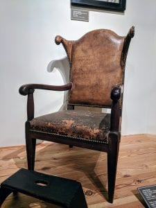 A photograph of an armchair displayed in the Dianne H. Furr Moravian Decorative Arts Gallery at the Frank L. Horton Museum Center. The armchair has dark wooden arms, legs, and front chair rail. The leather on the seat appears dark and worn. The leather on the backrest appears lighter in color than that on the seat. Small brass tacks hold the leather to the wood.