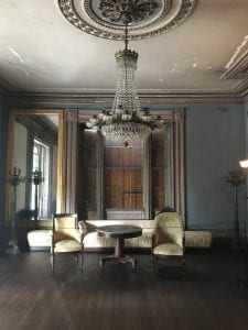 A large, dark room with a crystal chandelier and walls painted in a chalky blue paint.  A few pieces of nineteenth-century furniture stand at the far side of the room.  The grey ceiling paint is chipping, revealing white plaster beneath.