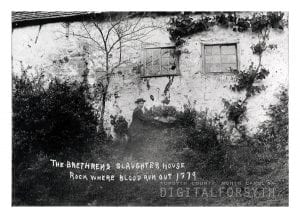 A man in a hat is photographed pointing at a drainage spout coming out of the old slaughterhouse in Salem. The man is standing outside of the building. Bushes and vines are overgrown and are covering part of the exterior.