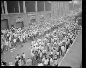 A large crowd standing outside of the “Bleacher Entrance” at Fenway Park. The crowd of people extends down Lansdowne Street. Most of the people are standing and a few Police Officers are riding horses.