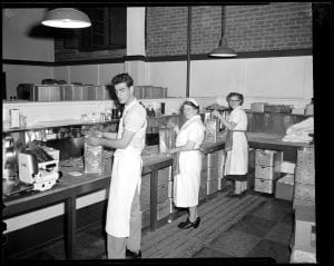 Three people in white aprons working in a kitchen at Fenway Park. Each of the individuals is holding a stack of hot dogs. The kitchen is large and industrial. Two of the individuals are female and one is male.