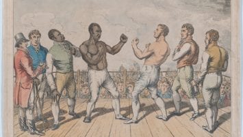 A hand-colored etching by George Cruikshank depicts the fight between Tom Molineaux, depicted on the left, and Tom Cribb, depicted on the right. They stand with fists raised ready to fight and are surrounded by five men in the ring. In the background a massive crowd come to watch the match is visible.
