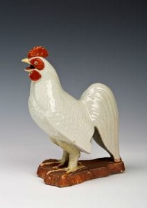 A white porcelain rooster, with red wattles and comb, standing on a painted brown based.