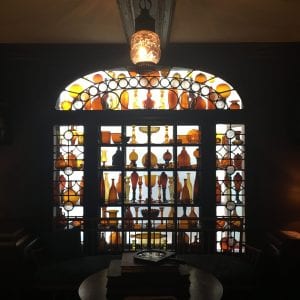 A glass lamp hangs above a large, back-lit window filled with seven shelves of bottles, candlesticks, and orbs made of matching, amber-colored glass.