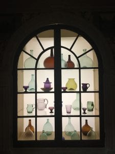 An arched window contains four shelves of multicolored glass objects, including green, purple, and brown cups, pitchers, and bottles, that are scattered in an intentional, but seemingly random, order.