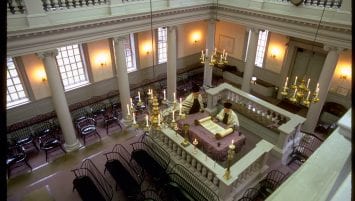 Interior image of Touro Synagogue looking down from the second floor. Bimah located in the center-right with two rabbis viewing a Torah