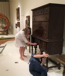 Two people study a piece of brown case furniture in a museum display. Karina Corrigan bends down to touch one of the drawers, while Greg Landry lies on the floor, studying the piece from below.