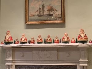 A close-up of a white mantle over a fireplace with a painting of a ship over it. On the mantle are twelve pair-shaped Santa figures with white beards, red coats, and black pants. They sit on false snow and between them are miniature pine trees.