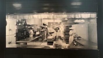A woman wearing a white cook’s hat frosts a cake in the middle of a 1930s professional bakery kitchen.