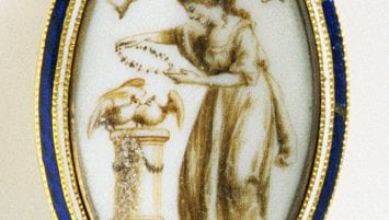 An almond shaped brooch with an image of a woman laying a wreath on two doves. Above the woman, a floating banner says “To Friendship.” The brooch has bands of white and blue enamel framing the image.