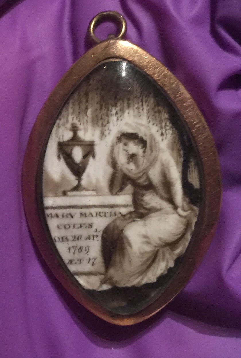 A pendant with a picture of a woman crying over a tomb with an urn on it.  The leaves of a weeping willow hang above her.  The tomb has an inscription with the name of the person who died and the date, 1789.
