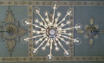 A chandelier seen from below. The branches form a star-like pattern. Behind, beige and coffee colored floral plasterwork can be seen against the blue of the ceiling.
