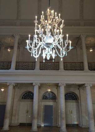A chandelier with many branches hangs in a large room.  Behind it is a walkway set on top of a series of limestone columns.  On the ground floor are three doors.