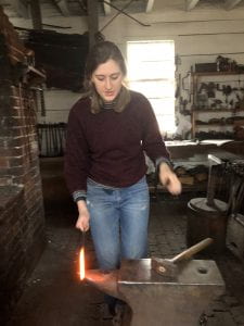 A young woman in blue jeans and a dark maroon sweater stands behind an anvil. She is holding a thin iron rod, the tip of which is glowing a bright whitish red color. She is about to pick up a hammer resting on the anvil.