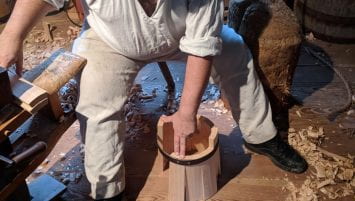 A man sits on a stool, holding together the staves of a bucket within an iron brace in one hand as he reaches for additional staves to fill the rest of the circle.