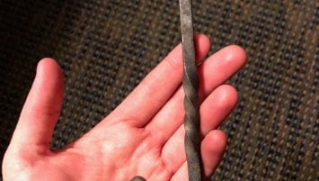 A left hand, palm facing up. Balanced across this hand is an S-hook, or iron bar shaped into an elongated S-shape. It has little curls at each end and a twist in the middle.