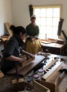 A woman uses a plane on a piece of wood in a workshop, while another woman in 18th-century garb looks on.