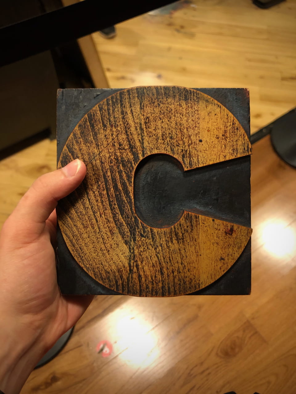 A left hand holding a square wood block cut to make the letter “C”. The person holding the block was also taking the picture. Previous ink use on this wood block accentuate the grain of the wood running at a slight diagonal across the “C.”