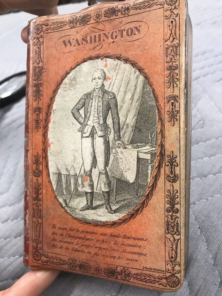 Glass flask covered with orange paper featuring a print of George Washington, the title “Washington,” and a poem in French.