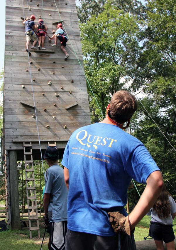 three students climb an outdoor rock wall while a Quest leader looks on from the ground