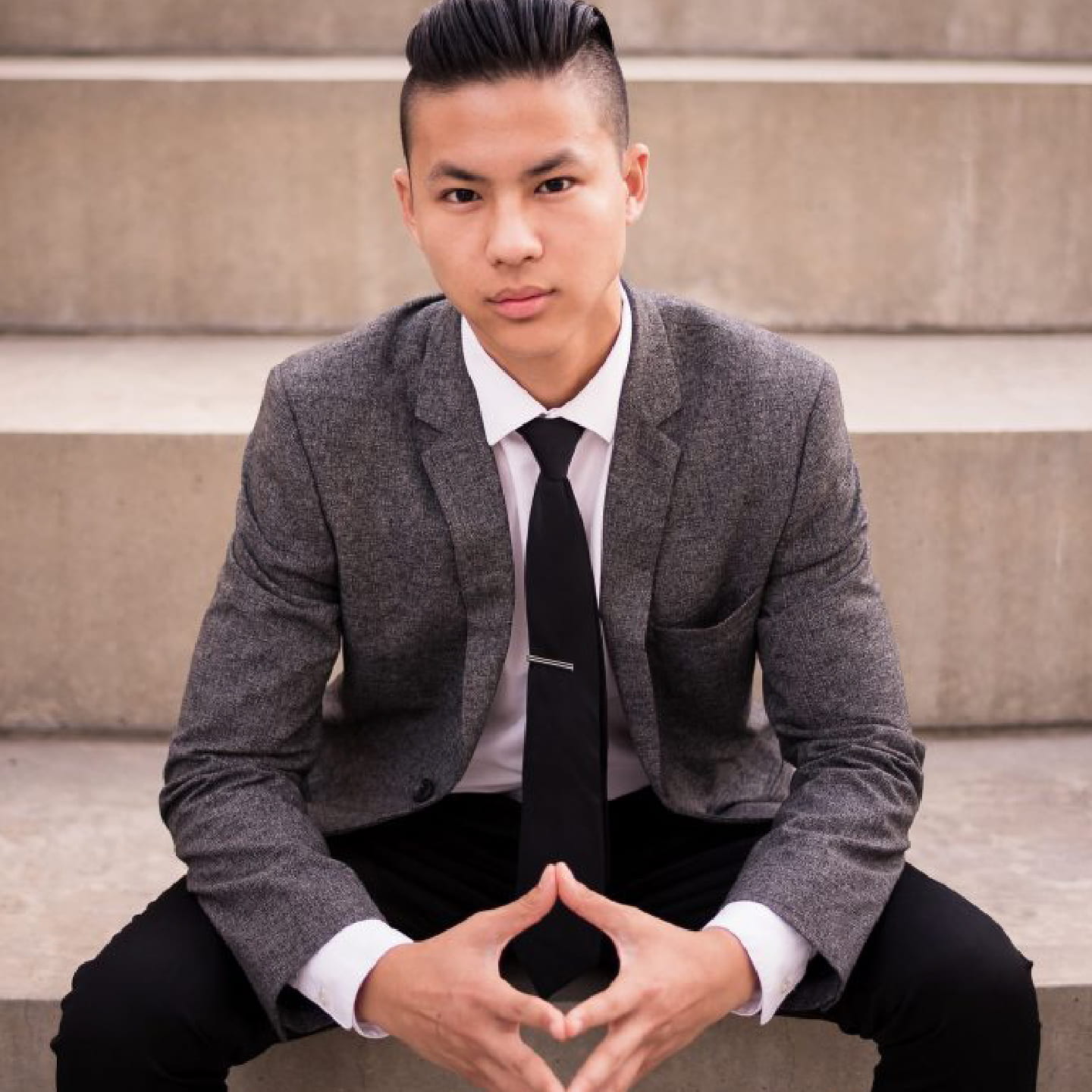 Kevin Li wears a gray blazer and black tie while sitting on concrete steps